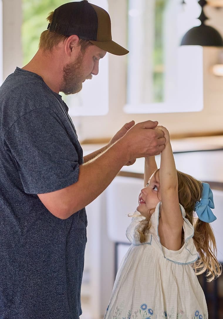 A father dances with his young daughter, holding her hands up, inside their home.