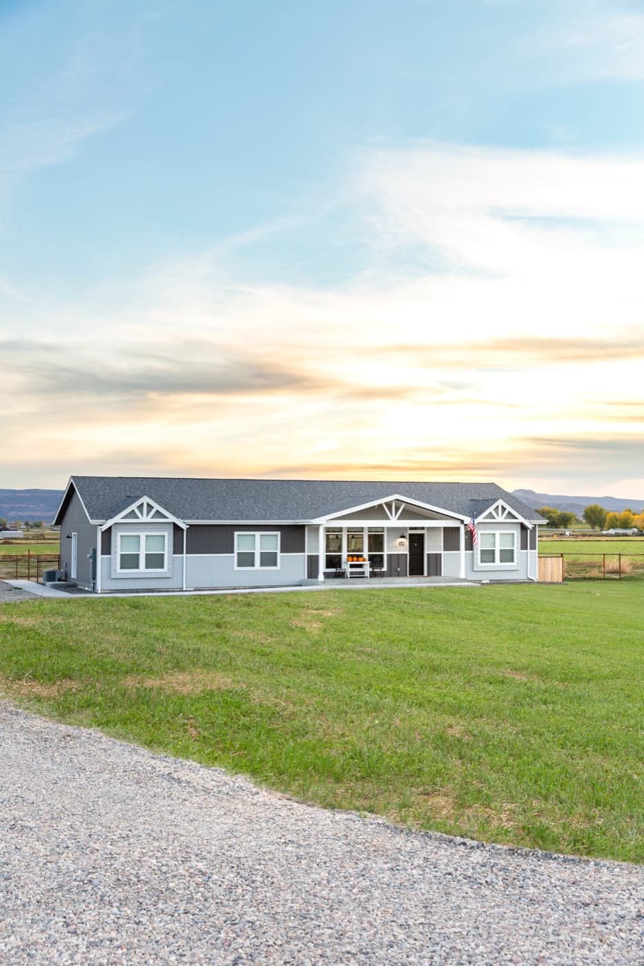 A single-story gray modern manufactured home with white trim on open rural land at sunset.