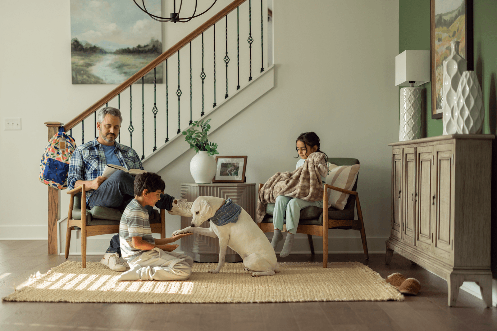 A family relaxing in their home's entryway, with a father reading, a boy petting the dog and a girl wrapped in a blanket.