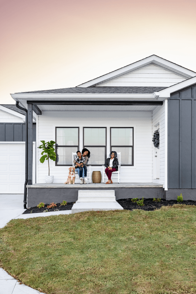 A family with their dog relaxing on the covered front porch of a white CrossMod home at dusk.