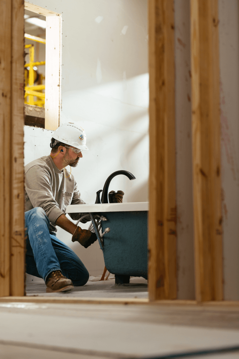 A worker in a hard hat installing a freestanding bathtub inside a framed home at a building facility.