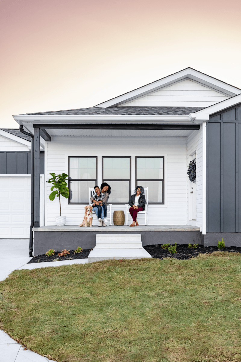 A family with their dog relaxing on the covered front porch of a white CrossMod home at dusk.