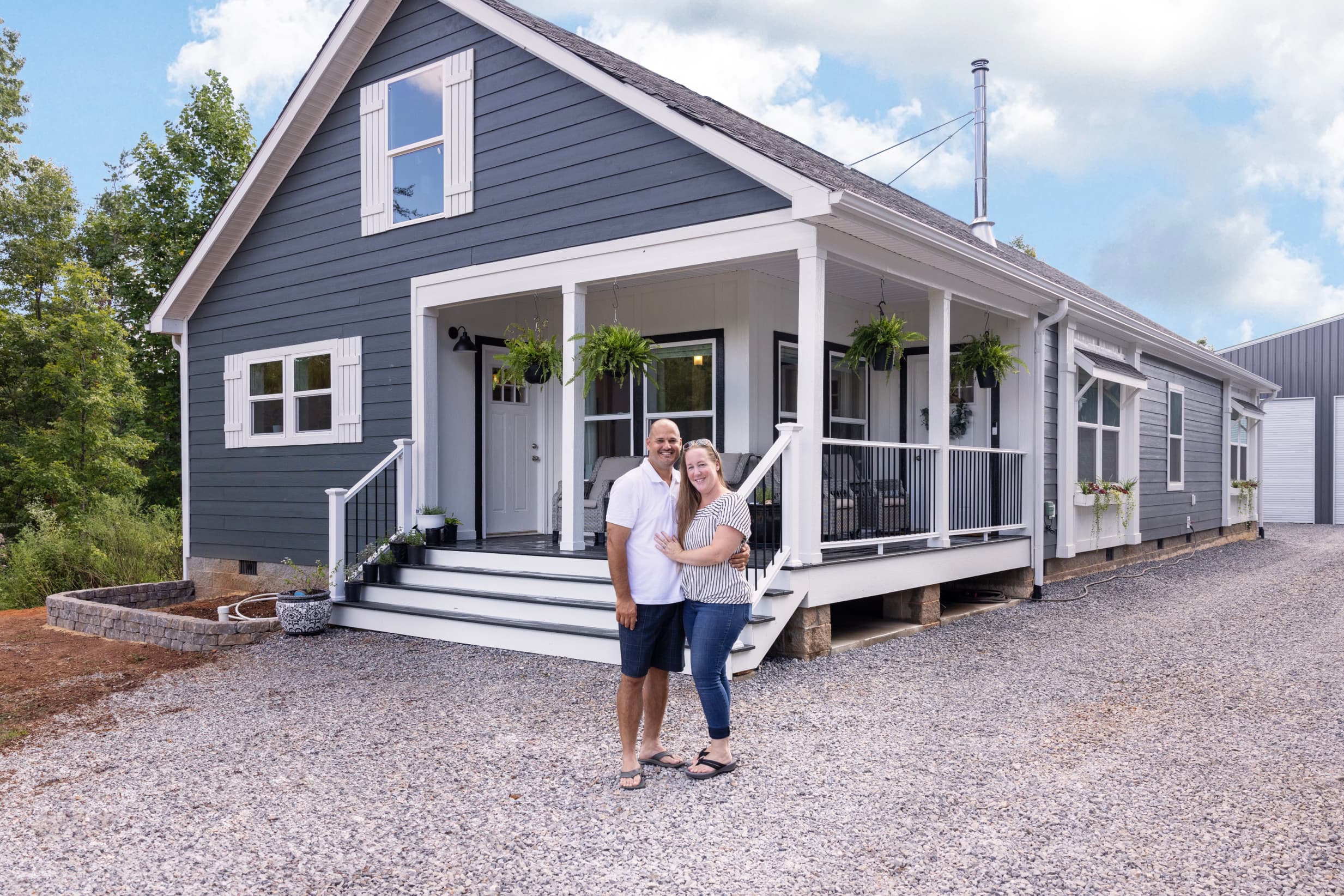 A smiling couple pose outside their front porch with ferns hanging and a blue exterior on their modern manufactured home.