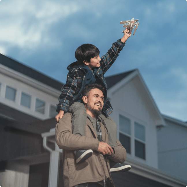A father carries his smiling young child on his shoulders in front of their home.