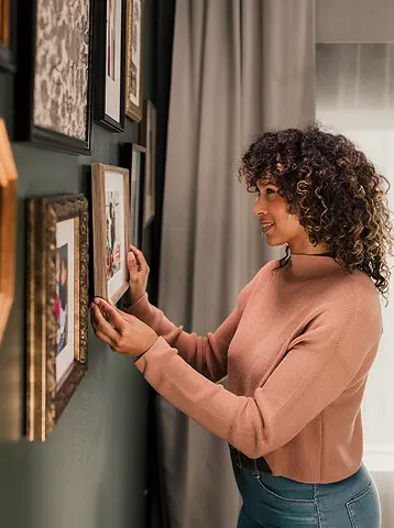 A woman hangs pictures in frames on a green wall inside her modern manufactured home.