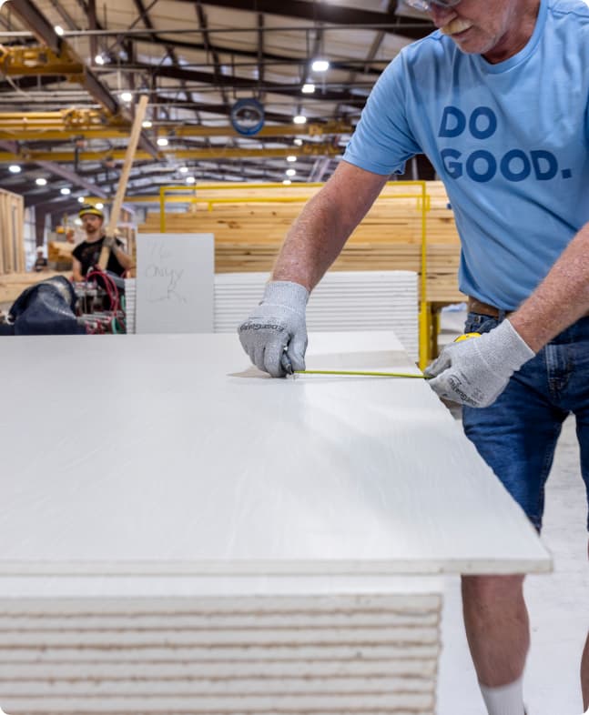 A builder works inside an indoor modern manufactured facility while measuring materials for a home.
