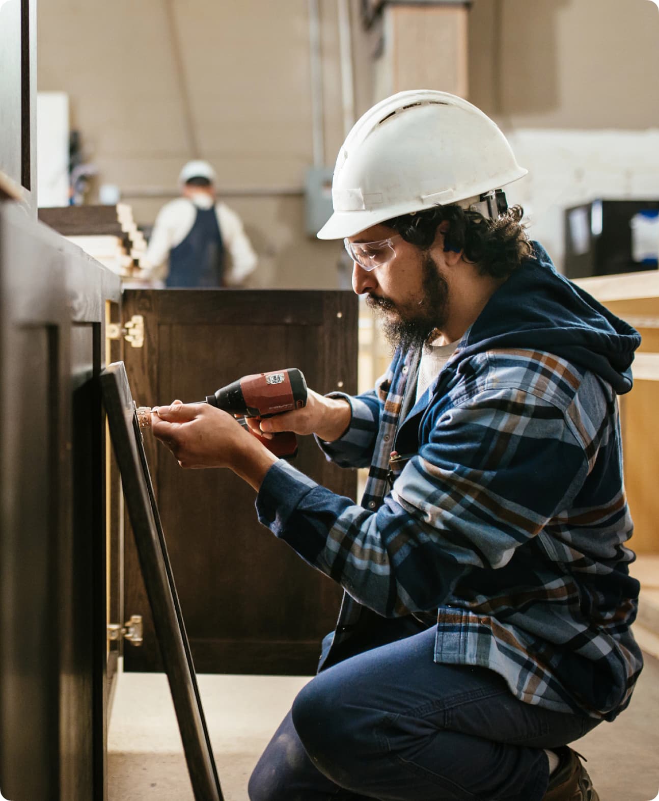 A man uses a power tool to mount a cabinet door to a kitchen cabinet while wearing a hard hat.