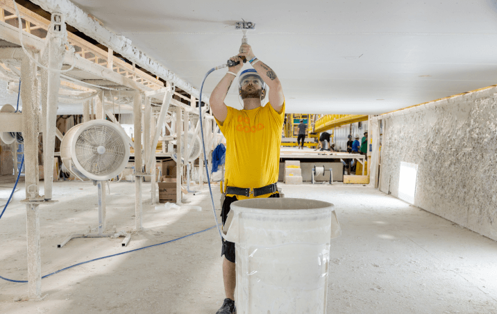 A worker at a building facility drills overhead beside a large clean collection bin.