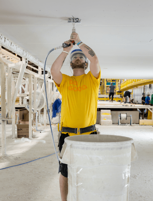 A worker at a building facility drills overhead beside a large clean collection bin.