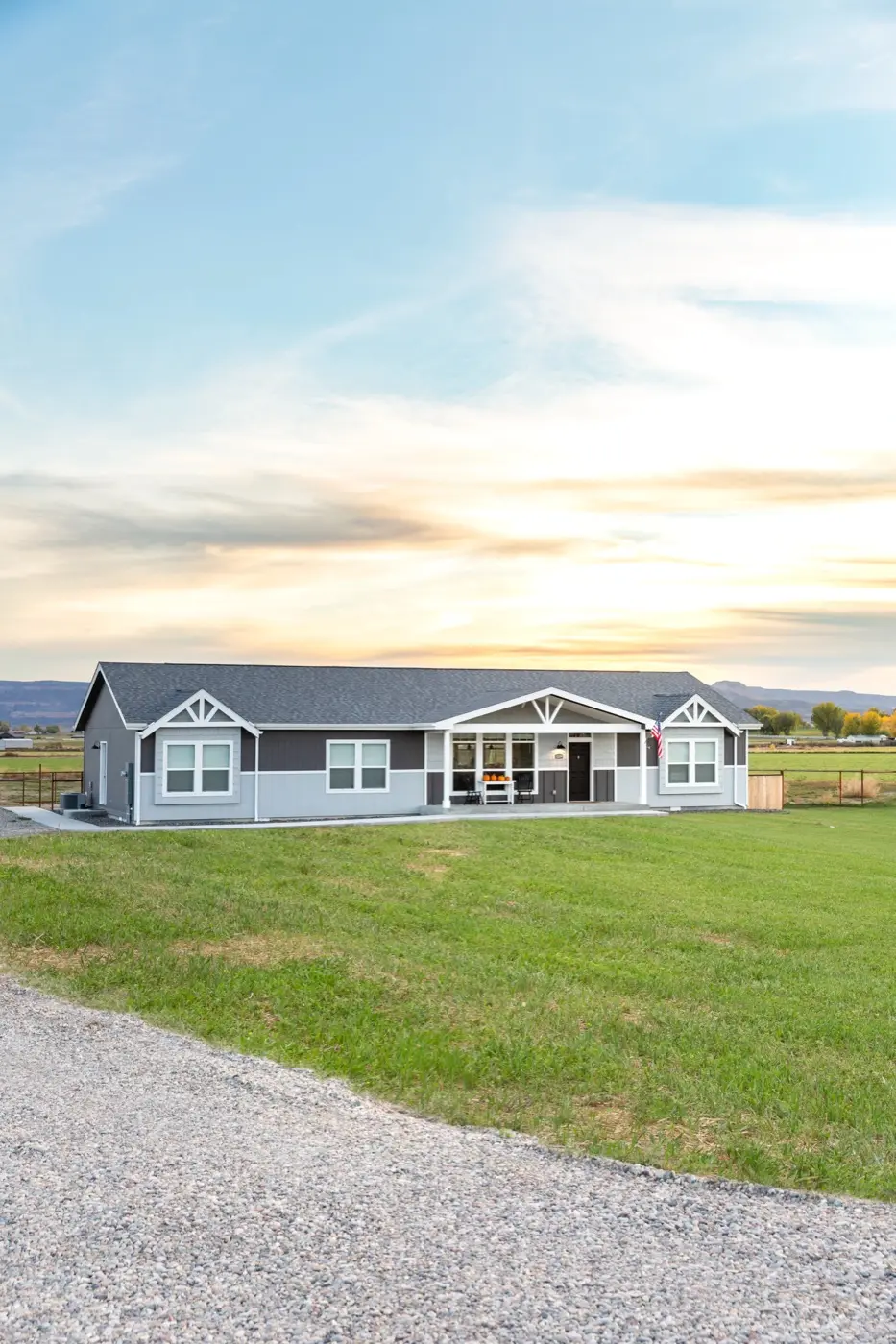 A single-story gray modern manufactured home with white trim on open rural land at sunset.